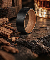 Black whiskey barrel wedding ring with natural wood interior displayed beside oak wood and a glass of whiskey