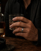 A close-up of a man's hand holding a whiskey glass with amber liquor, wearing a simple matte silver band ring on his ring finger. A dark bottle and wooden bar surface are visible in the dimly lit background.
