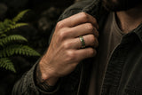 Close-up lifestyle photograph of a man wearing a silver tungsten wedding ring with green moss agate inlay in a dark cinematic outdoor editorial setting.