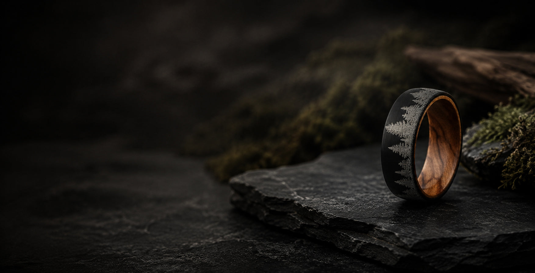 Luxury men’s black woodland wedding ring with engraved tree pattern positioned in a rugged natural-inspired hero banner with dark slate textures and cinematic lighting.