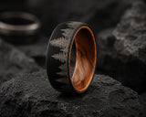 Macro close-up of a black men’s wedding ring with engraved forest pattern and natural wood sleeve resting on dark textured volcanic stone.