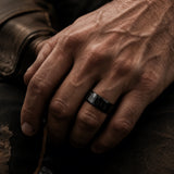 Close-up of a man’s hand wearing a black wedding band against rugged dark clothing with moody cinematic lighting.