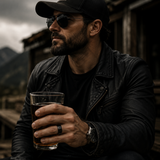 Man wearing sunglasses and a black leather jacket holding a whiskey glass while wearing a black wedding band outdoors.