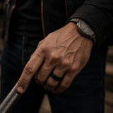Close-up of a rugged male hand wearing a black ring and watch while gripping a metal bar in a cinematic outdoor setting.