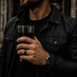Man in a black leather jacket holding a whiskey glass while wearing a black wedding ring and minimalist watch.
