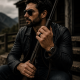 Rugged man wearing sunglasses and a black wedding band while holding a rifle outside a rustic cabin in the mountains.
