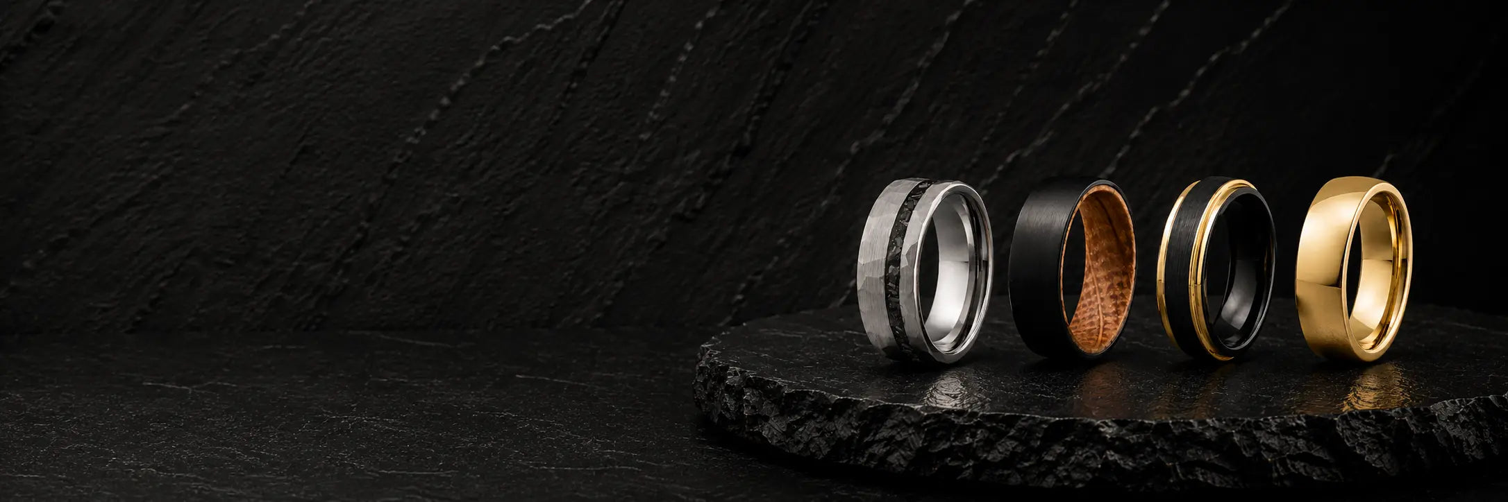 Men's wedding ring collection showcase featuring whiskey barrel wood, meteorite, gemstone and hammered tungsten wedding rings on a dark luxury background.