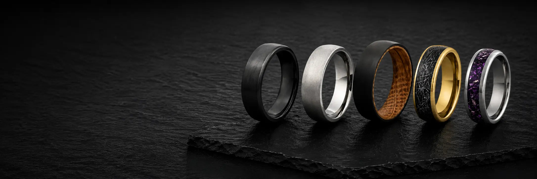 Men's wedding ring materials collection hero image featuring titanium, whiskey barrel wood, meteorite and gemstone wedding rings displayed on a dark slate background with space for text overlays on the left.