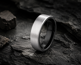 Close-up of brushed silver tungsten wedding ring with black interior resting on dark stone showing durable textured finish