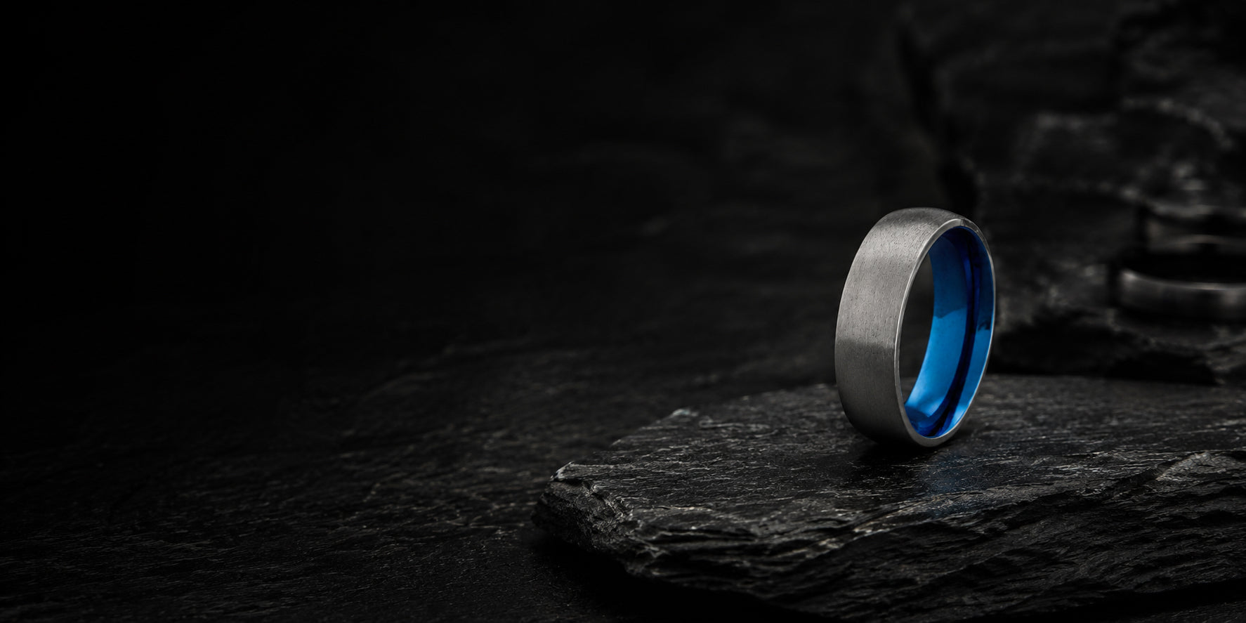 Luxury desktop hero banner featuring a silver and blue titanium wedding ring on dark charcoal slate textures