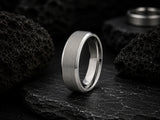 Silver brushed titanium men’s wedding ring on dark volcanic rock with cinematic moody lighting and rugged charcoal slate background.