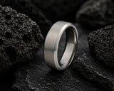 Macro close-up of a brushed silver titanium men’s wedding ring resting on dark volcanic rock with dramatic luxury lighting.
