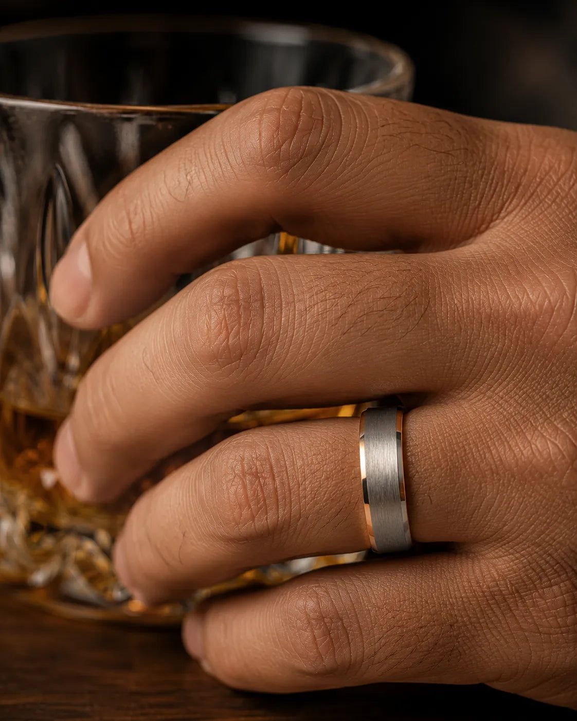 Macro close up of a silver brushed tungsten wedding ring with polished beveled edges and a whiskey barrel wood interior worn while holding a crystal whiskey glass.
