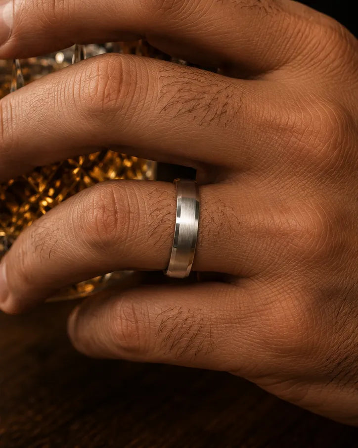 Macro close up of a silver beveled whiskey barrel wood wedding ring worn on a man's finger while holding a crystal whiskey tumbler.