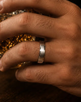 Macro close up of a silver beveled whiskey barrel wood wedding ring worn on a man's finger while holding a crystal whiskey tumbler.