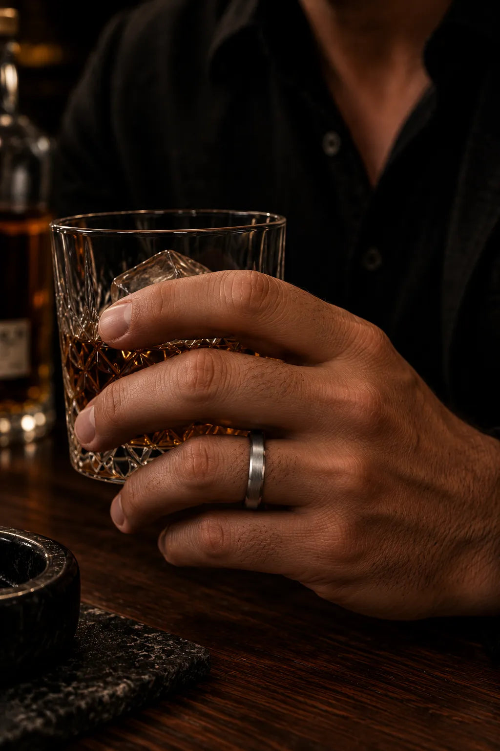 Man wearing a silver beveled whiskey barrel wood wedding ring while holding a crystal glass of whiskey in a dark cinematic bar setting.