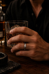 Man wearing a silver beveled whiskey barrel wood wedding ring while holding a crystal glass of whiskey in a dark cinematic bar setting.
