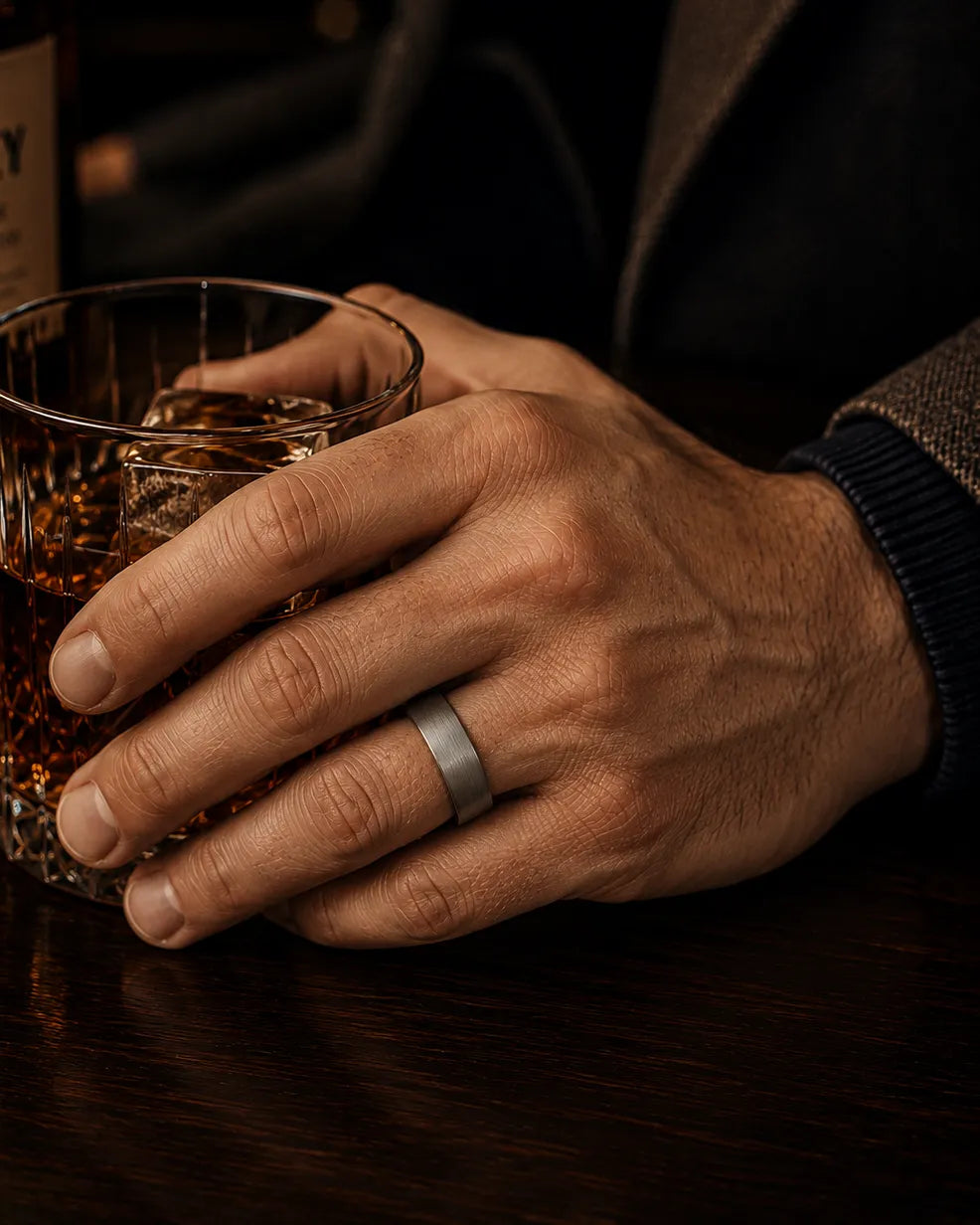 Man wearing a silver tungsten wedding ring with a brushed finish and whiskey wood interior while holding a whiskey glass in a dark luxury bar.