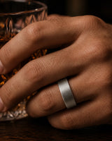 Close-up macro photograph of a silver tungsten wedding ring with a brushed finish and whiskey wood interior worn while holding a crystal whiskey tumbler in warm cinematic lighting.