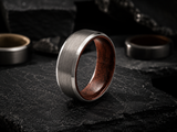 Brushed silver men’s wedding ring with dark whiskey barrel wood interior resting on volcanic slate in a moody cinematic setting