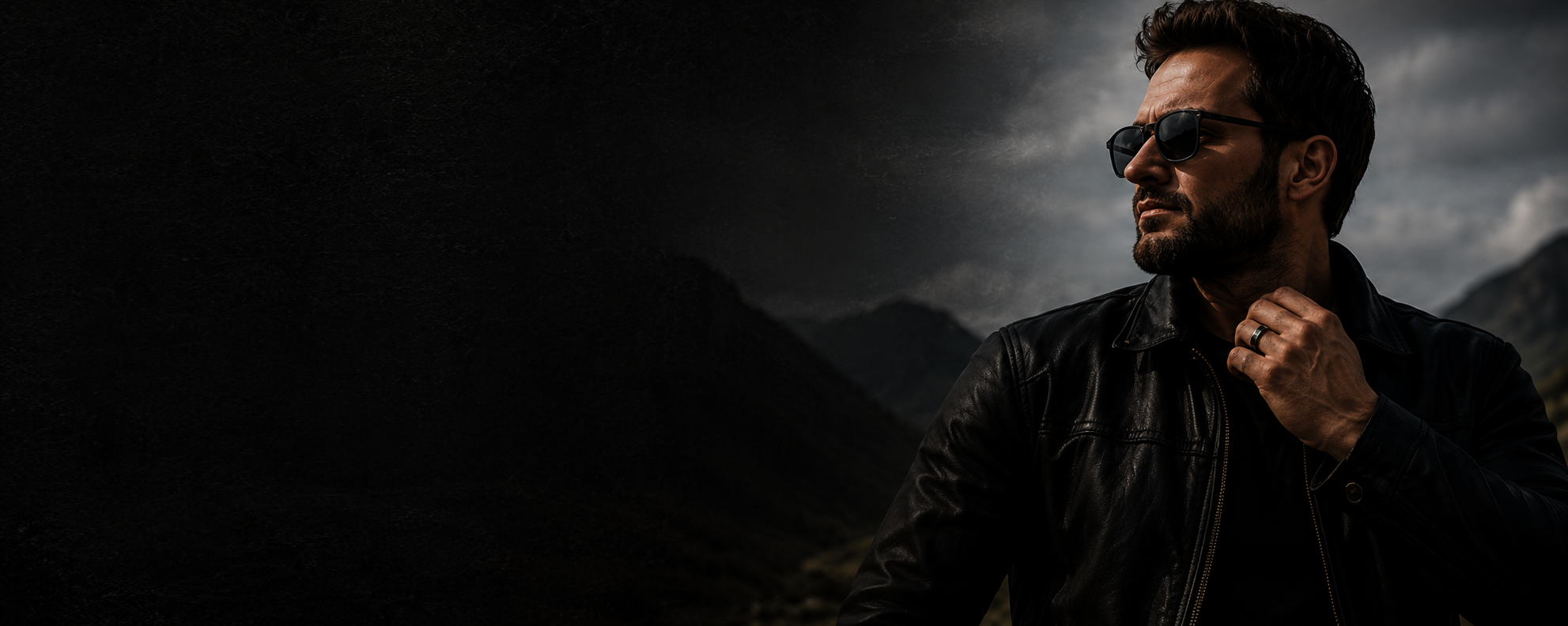 Moody cinematic portrait of a man wearing a black wedding band and leather jacket against a dark mountain landscape with stormy skies and rugged masculine styling.